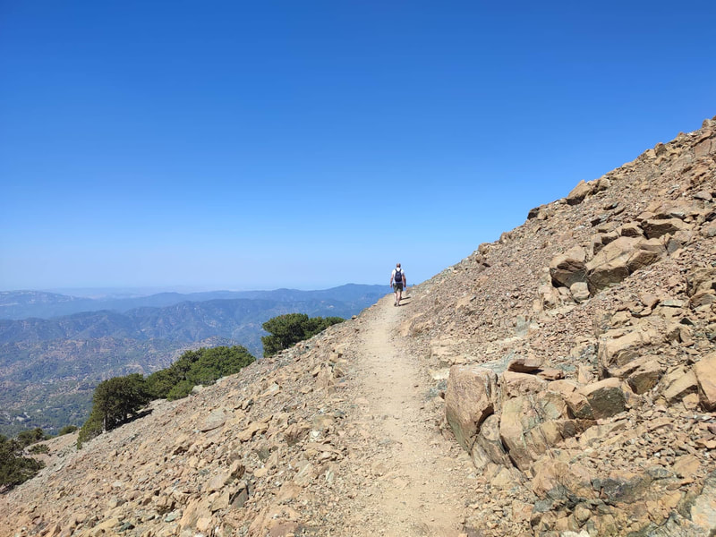 A walking trail on a steep slope of a rocky peak under a blue sky. A walker in the background.