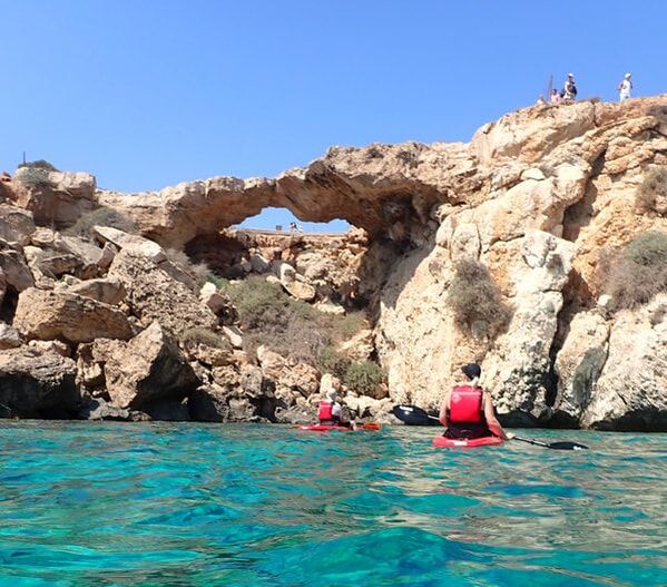 Pictureseeing the back of two kayakers in single red siton top kayaks in crystal blue sea. A rocky arch in the background