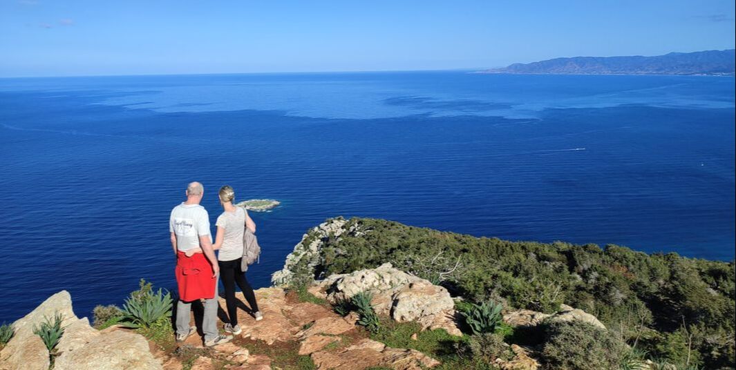 a man and a woman in short sleeves on a rocky peak, blue sky and blue calm sea in the background